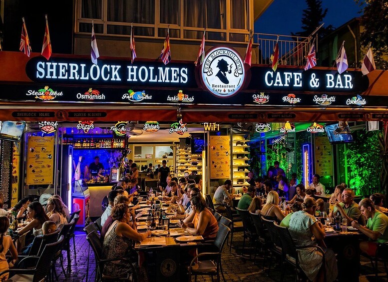 A busy outdoor restaurant named 'Sherlock Holmes Cafe & Bar' with various food and drink options. 4