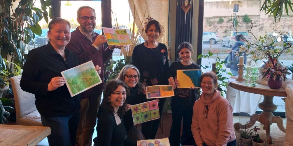 A group of people holding colorful artwork in an indoor setting with plants and a window. 3