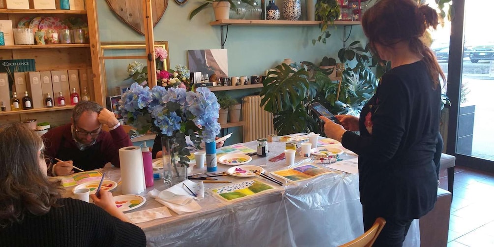 A painting class with participants seated at a table covered with art supplies and canvases. 1