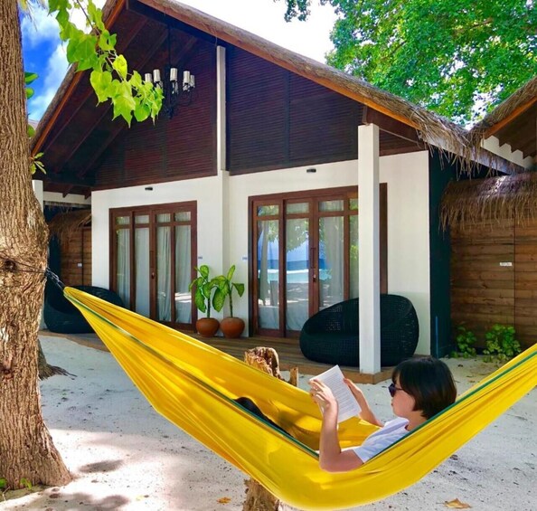 A person reading in a yellow hammock outside a wooden house with a thatched roof. 2