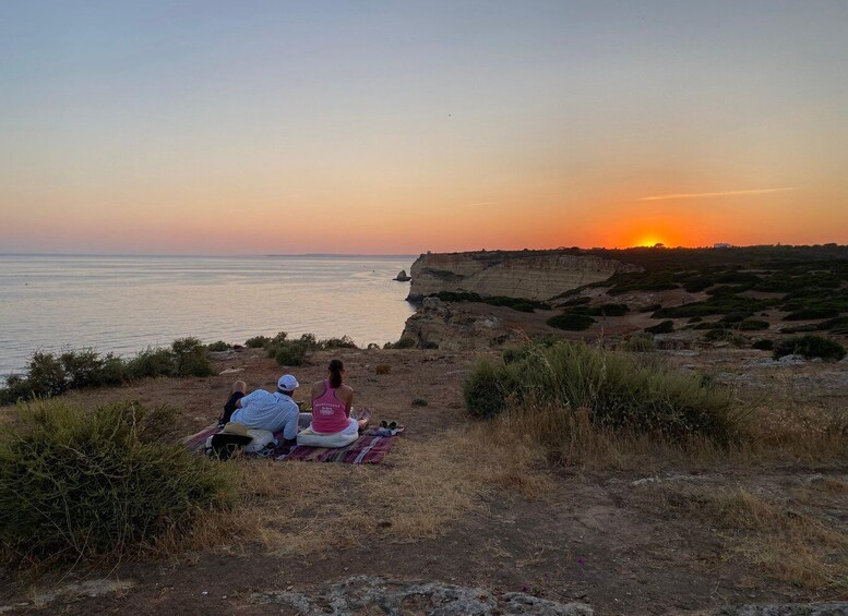 A sunset over a coastal landscape with two people sitting on a blanket. 1