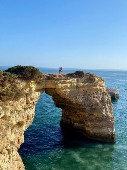 A natural rock arch formation over a body of water with two people standing at the top. 2