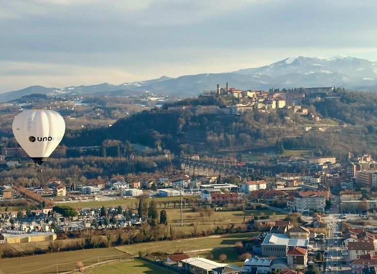A hot air balloon with 'unoene' branding flying over a town with a bridge and mountains in the background. 3