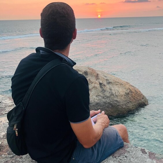 A man sitting on a rock by the ocean during sunset. 2