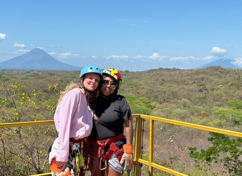 Two people wearing helmets and harnesses, standing on a yellow railing with a mountainous landscape in the background. 5