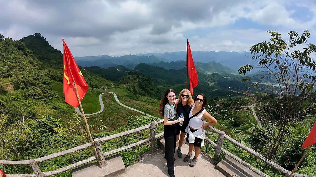 A group of people standing on a concrete platform with red flags, overlooking a winding road and lush green hills. 1
