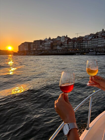 Two people holding wine glasses on a boat with a sunset and buildings in the background. 1