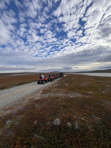 A group of people on ATVs driving on a gravel path through a tundra landscape with sparse vegetation and a cloudy sky. 2