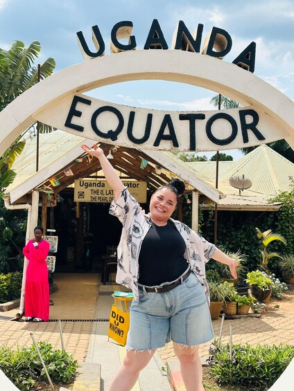 A person standing in front of a sign that reads 'UGANDA EQUATOR' with a tropical garden in the background. 1