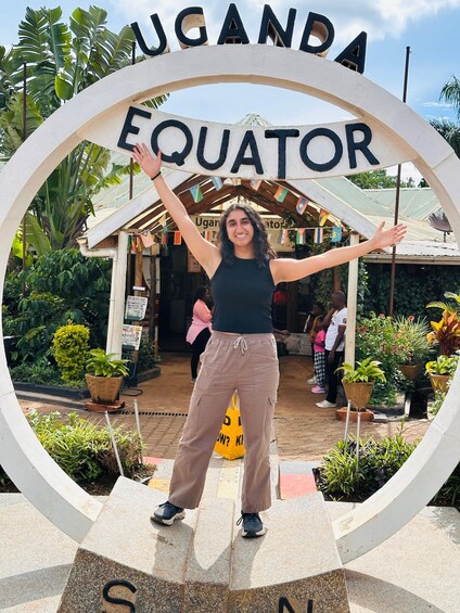 A person standing in front of a sign that reads 'UGANDA EQUATOR' with arms raised. 5