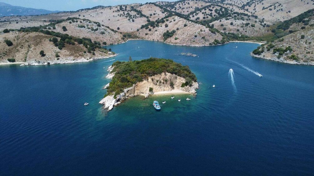A small island surrounded by clear blue waters, with boats anchored near the shore and a wake trail indicating recent boat activity. 1