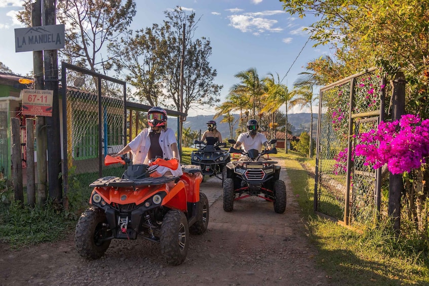 Three people on ATVs on a dirt path with a sign reading 'LA MANUELA' and a fence with a gate. 1