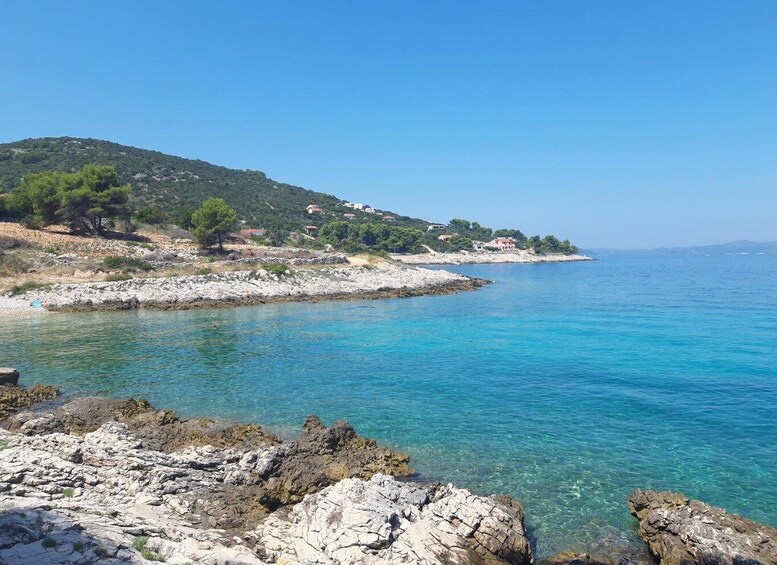 A coastal landscape with clear turquoise waters, rocky shoreline, and a hilly backdrop with scattered buildings. 5