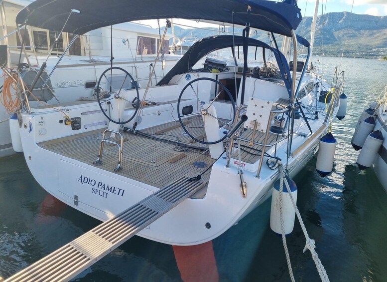 A white sailboat named 'ADIO PAMETI' docked at a marina with a mountainous backdrop. 3