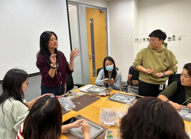 A group of people in a meeting room with a whiteboard, a table with jewelry-making materials, and a door in the background. 3