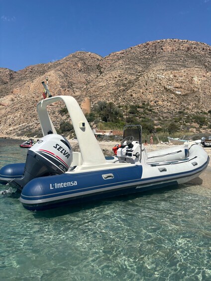 A blue and white inflatable boat named L'Intensa with a SELVA motor, docked near a rocky shoreline with clear water. 2