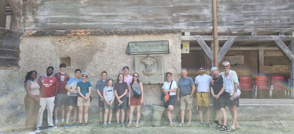 Un groupe de personnes se tient devant un bâtiment rustique, doté d’un mur en pierre et d’un toit en bois. 2