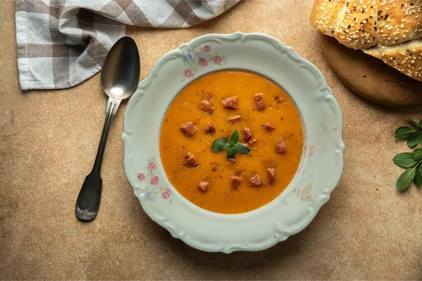 A bowl of soup with croutons and a sprig of herb, a spoon, and a checkered cloth on a textured surface. 2