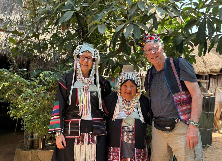 Three people in traditional attire standing in front of a thatched structure. 1