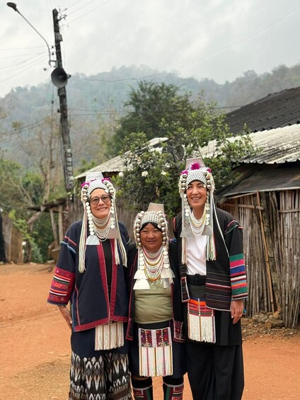 Three individuals in traditional attire with elaborate headdresses and jewelry, standing in front of a rustic building with a dirt path and vegetation in the background. 3