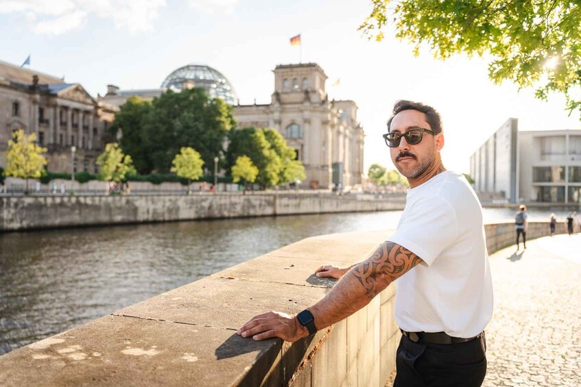 A man with a tattoo leaning on a bridge railing with a river and buildings in the background. 4