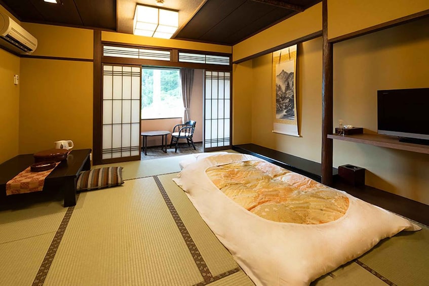 A traditional Japanese room with tatami flooring, a futon bed, a television, and a sliding door leading to a balcony. 1
