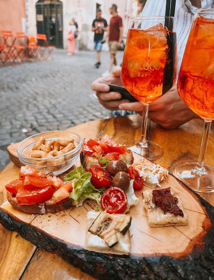 A wooden board with various foods and two glasses of a red beverage, possibly a cocktail, on a cobblestone street. 5