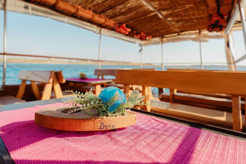 A wooden table with a blue decorative item and greenery on a pink mat, a wooden bench, and a thatched roof structure on a boat. 5