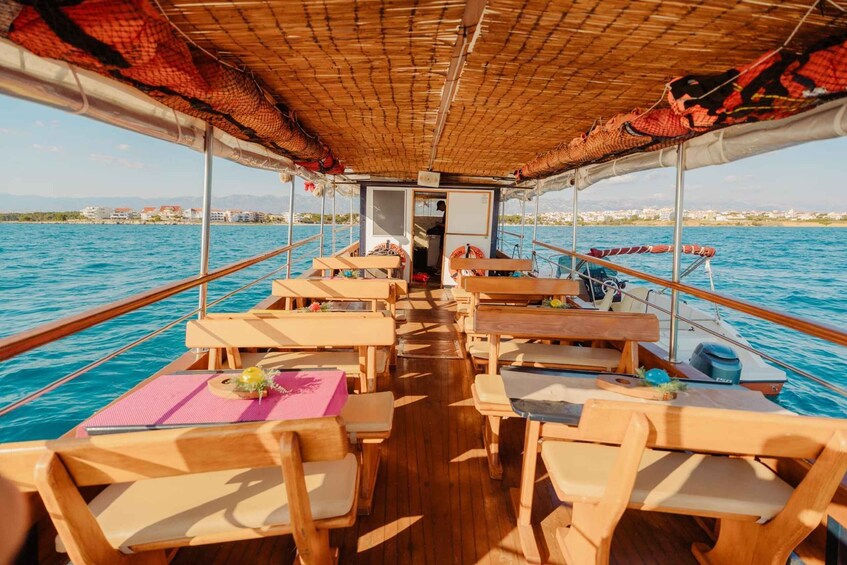 A boat with wooden tables and chairs, a covered area, and a view of the sea and buildings in the background. 3