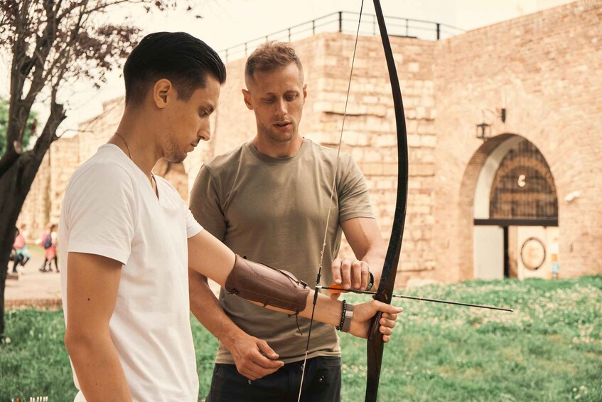 Two individuals practicing archery outdoors with a historical building in the background. 3