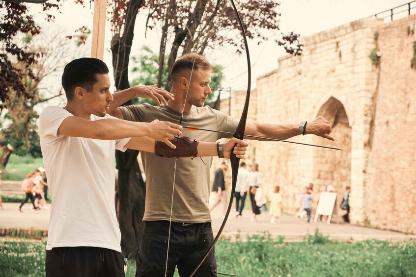 Two people practicing archery outdoors with a stone wall and trees in the background. 4