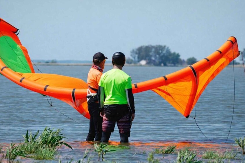 Two people standing in shallow water with a large orange kite. 1