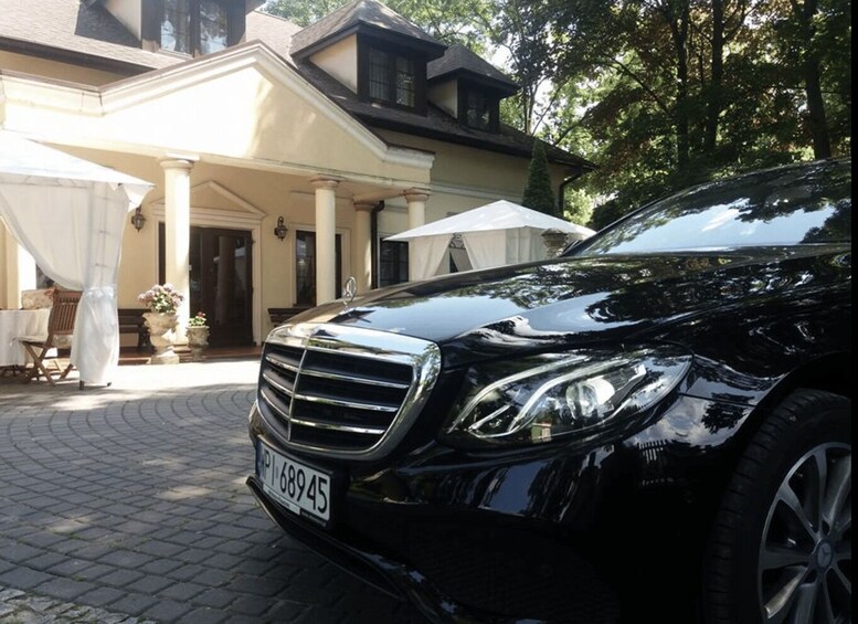 Musta Mercedes-Benz oli pysäköitynä valkoisen rakennuksen eteen. Rakennuksessa oli pylväitä ja katettu terassi. 5