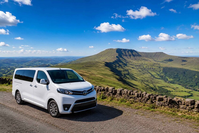 A white van parked on a gravel path with a scenic view of rolling hills and a mountain in the background. 1