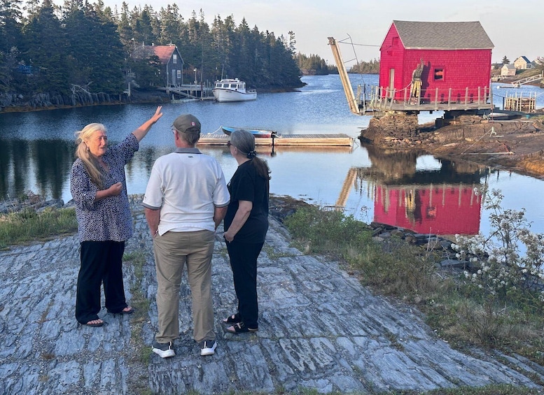Three people standing on a rocky shore, a red building, and a boat on the water. 1