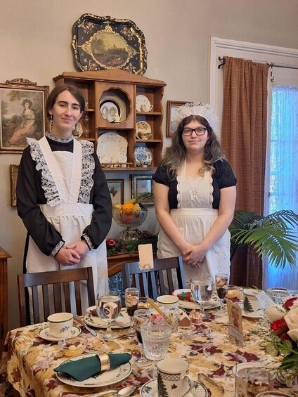 Two individuals in historical attire standing at a table set with fine china, glassware, and a floral tablecloth. 4