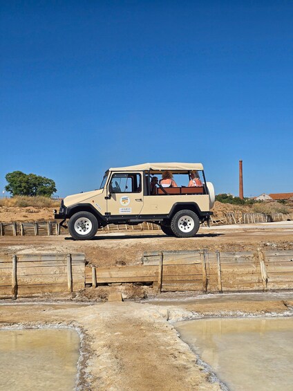 A beige off-road vehicle with passengers, parked near a wooden fence and a salt flat with water pools. 1