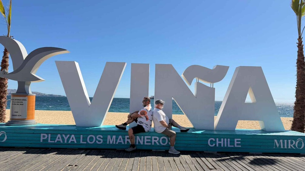 A wooden boardwalk with large white letters spelling 'VINA' and 'CHILE' along with palm trees and a clear blue sky. 1
