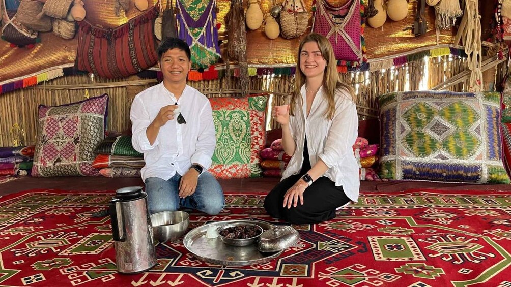 Two people seated on a patterned carpet with traditional textiles and pottery in the background. 2