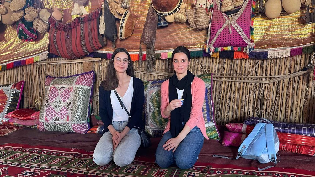 Two people sitting on a patterned rug with a backdrop of woven textiles and decorative pillows. 4