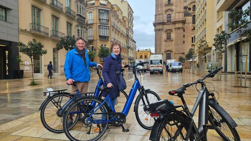 Two people standing next to bicycles in a city square with historic buildings in the background. 2