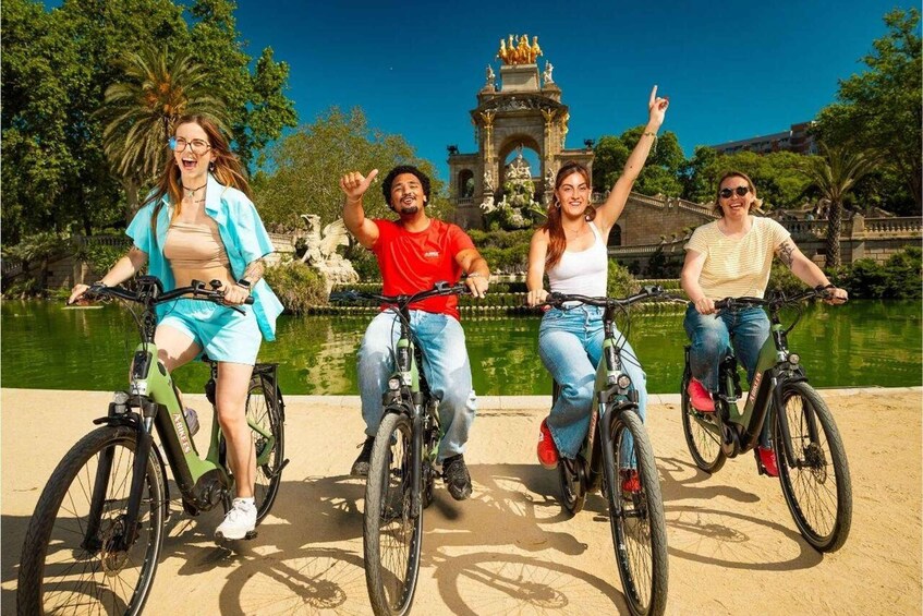 Four people riding bicycles on a paved path with a water feature and a fountain in the background. 1