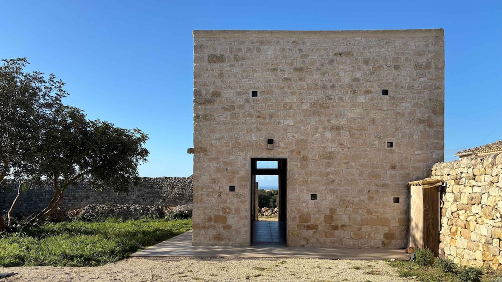 A stone building with a central doorway, surrounded by greenery and a clear sky. 1