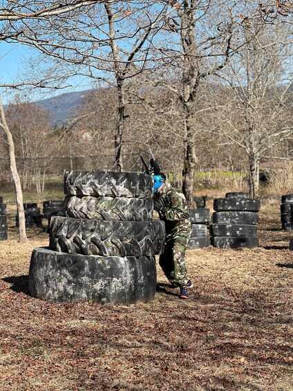 A person in camouflage gear standing next to a stack of tires in a wooded area with leafless trees. 2