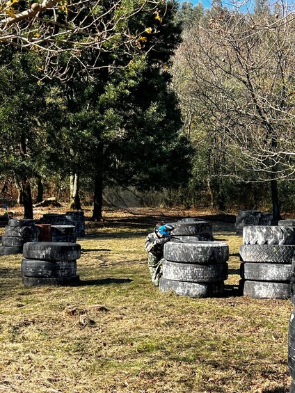 A person in camouflage gear aiming a paintball gun at a target made of stacked tires in a wooded area. 4