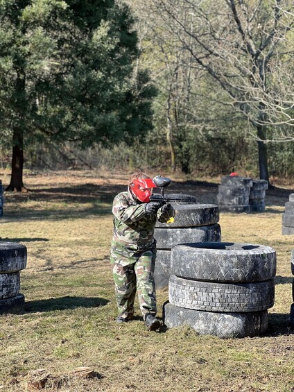 A person in camouflage gear aiming a paintball gun at a target made of stacked tires in an outdoor setting with trees in the background. 5