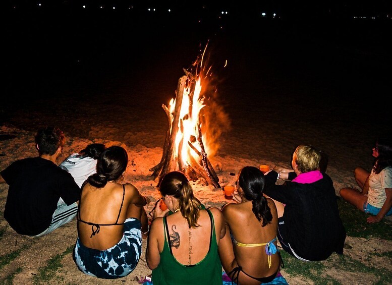 A group of people sitting around a campfire on a beach at night. 1