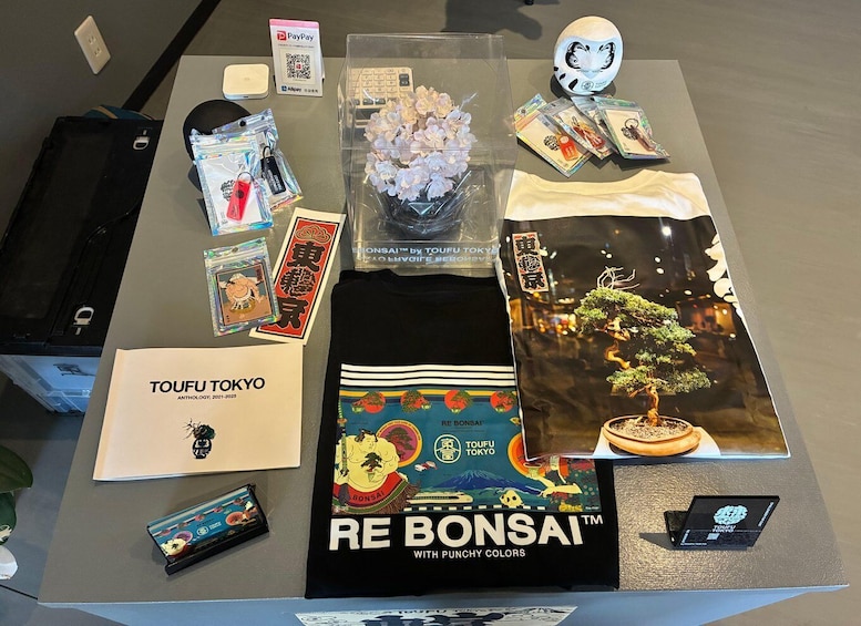 A table with various items including a book titled 'TOFU TOKYO,' a black t-shirt with 'RE BONSAI' print, a bonsai tree, and some cards. 2