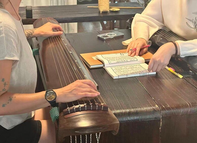 Two people playing a stringed musical instrument, with a book and a pen on the table. 2