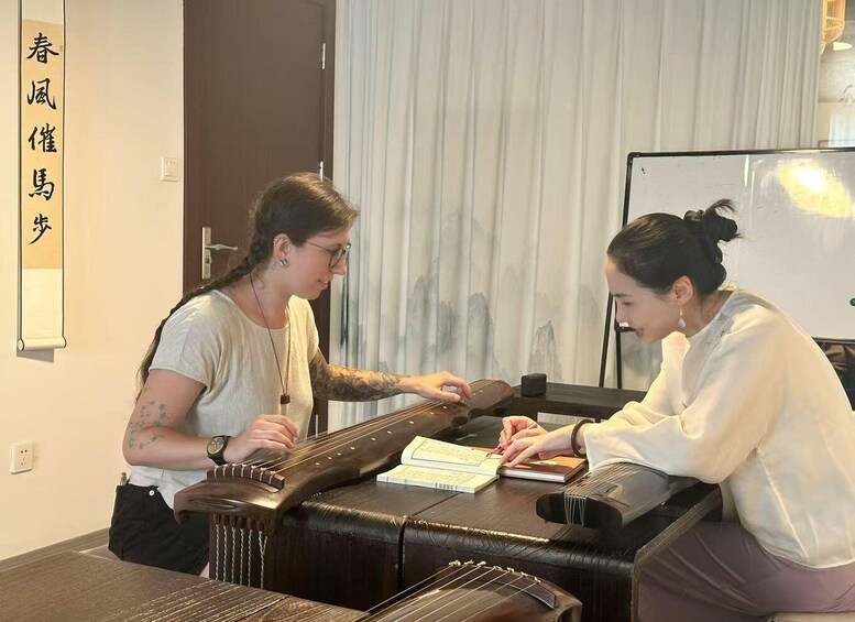 Two people playing a traditional stringed instrument, possibly a guzheng, with sheet music on the table. 1